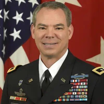 Bert Mizusawa in Army dress uniform with service ribbons, in front of a United States flag and a red flag.