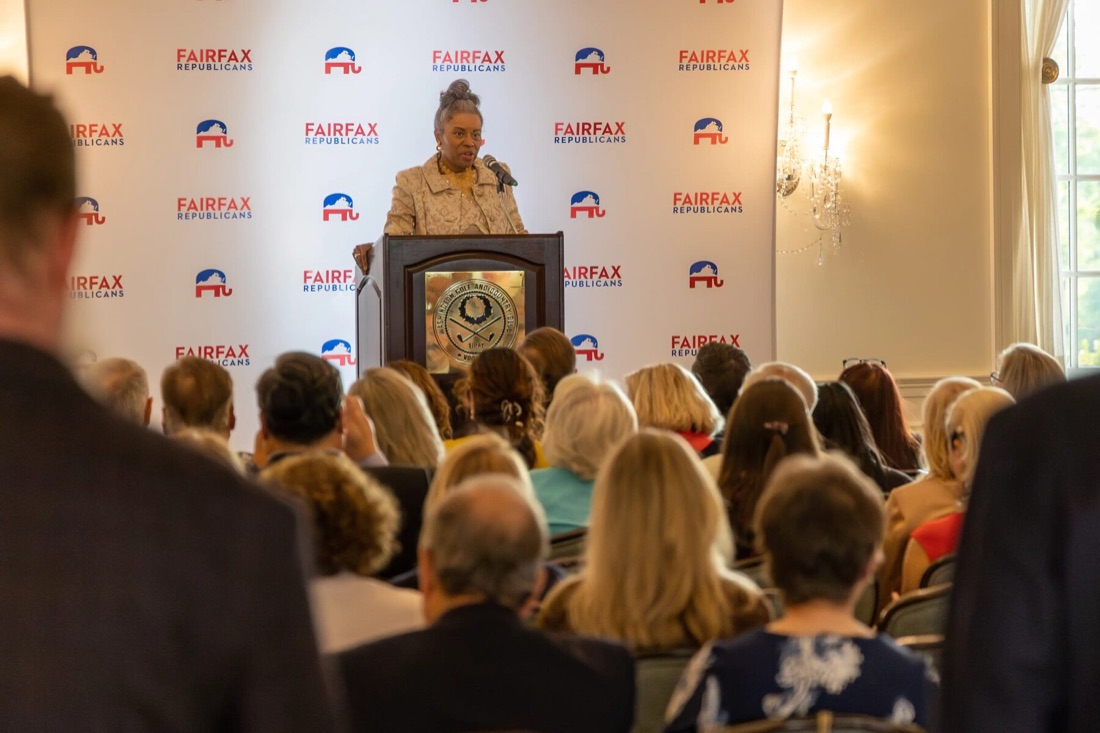 Winsome Earle-Sears speaks at a podium beneath the Fairfax Republicans step-and-repeat to a full seated audience in a chandelier-lit ballroom.