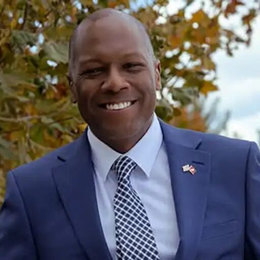 David Williams in a blue suit and patterned tie with a flag pin, outdoors against autumn foliage.
