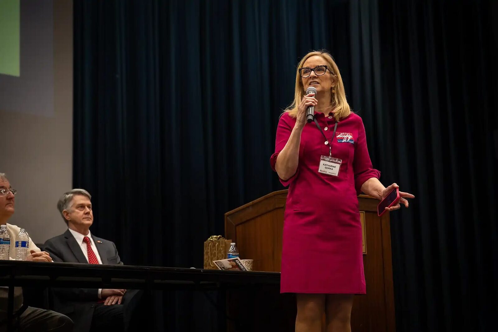 Chairwoman Katie Gorka, in a magenta dress and wearing her convention name badge, speaks into a handheld microphone on a dark stage; two men are seated at a table behind her.