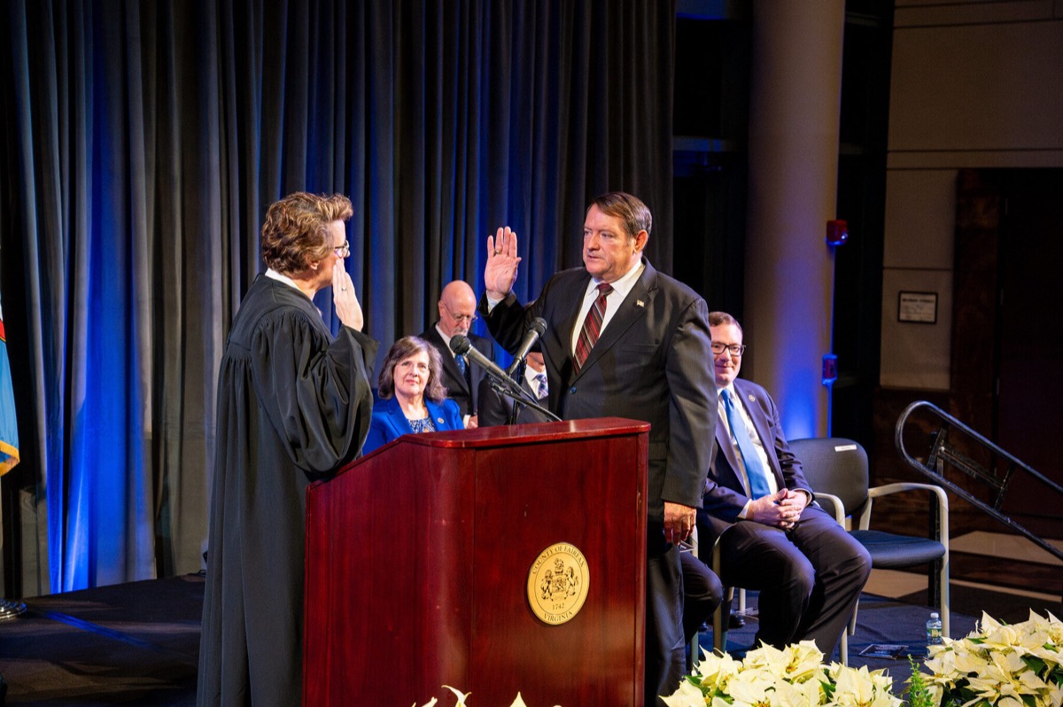 Supervisor Pat Herrity raises his right hand as a judge in black robes administers the oath of office, with officials seated against a blue drape.