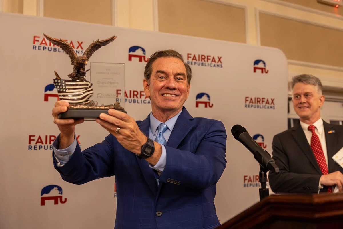 Chris Plante in a blue suit holds the Freedom Award eagle trophy aloft, laughing, in front of a Fairfax Republicans step-and-repeat while a second man applauds at the podium.