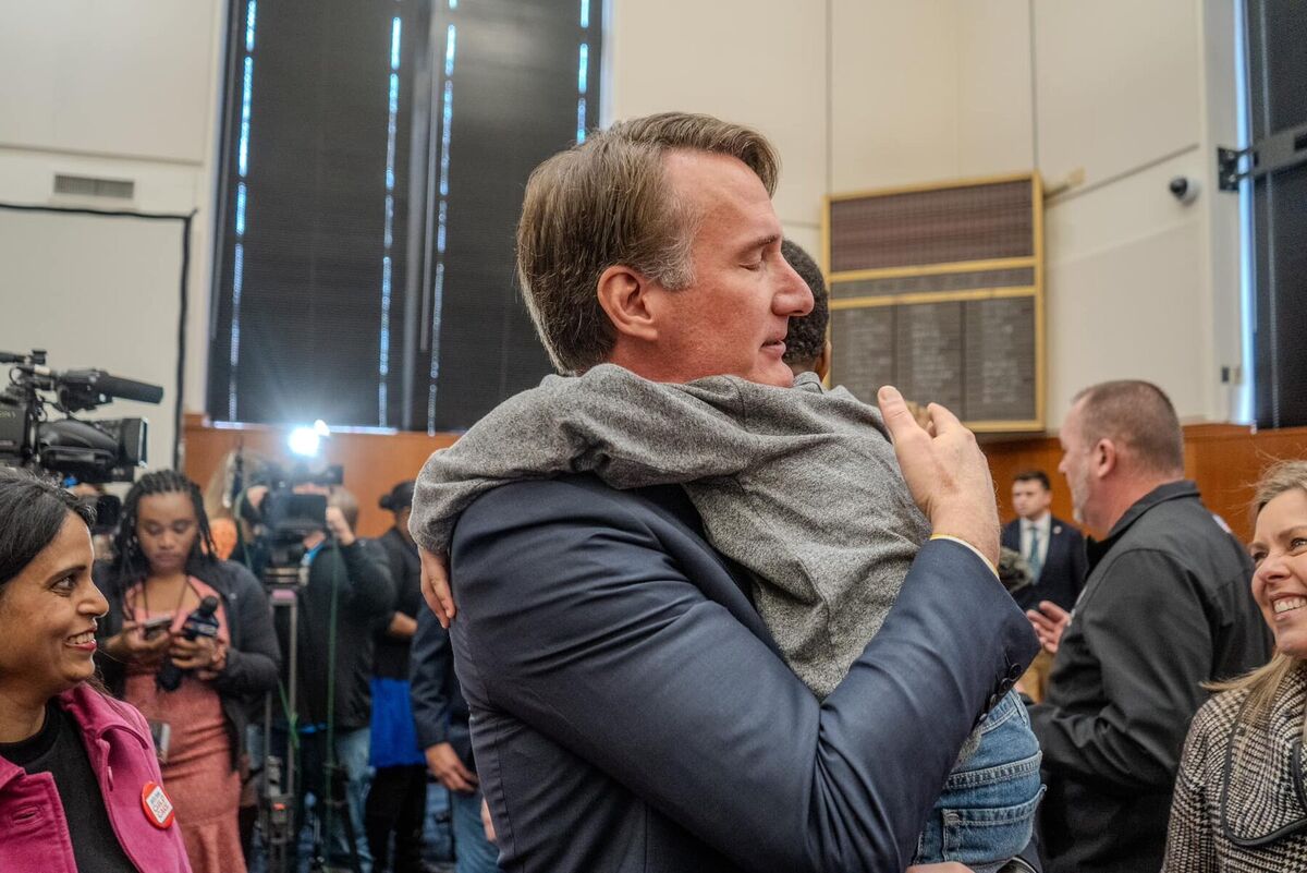 Glenn Youngkin lifts a young supporter in a hug in a crowded room as press cameras look on.